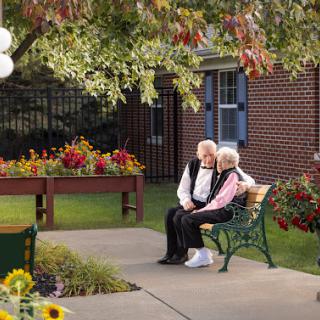 An elderly couple enjoys a serene moment together on a bench surrounded by colorful flowers in an autumn setting.