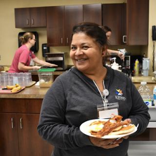 A joyful person holding a plate of bacon and pancakes in a busy kitchen.