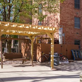 A sunny patio area with a wooden pergola, bench, and bicycles near a brick building.
