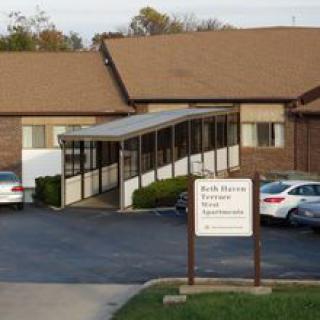 A small brick building with a parking lot and a sign in front, featuring cars parked near the entrance.