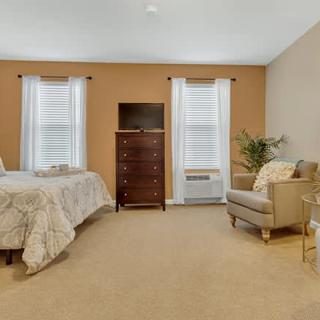 A cozy bedroom with a neatly made bed, a dresser, an armchair, and two windows with white curtains.