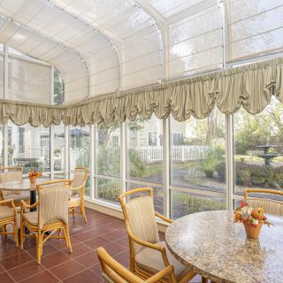 A bright sunroom with glass walls, featuring a dining area with tables and chairs under abundant natural light.