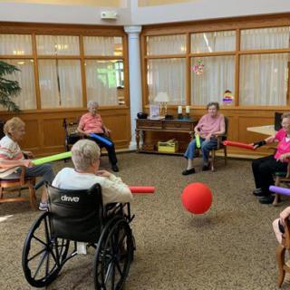 A group of seniors participating in a fun indoor ball game using noodles while seated in a circle.