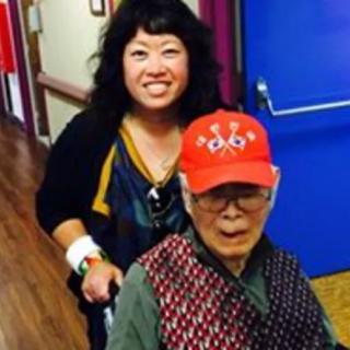 A smiling woman stands behind an elderly man in a wheelchair indoors.