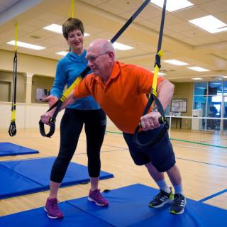 A senior man performs TRX exercises with the guidance of a trainer in a gym setting.
