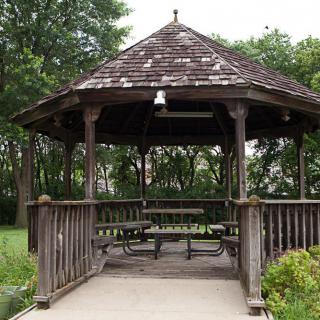 A wooden gazebo with picnic tables, surrounded by greenery in a park setting.