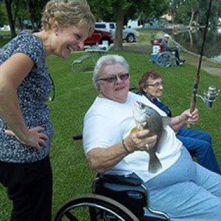 A person in a wheelchair proudly holding a fish they caught, surrounded by others enjoying a day outdoors.