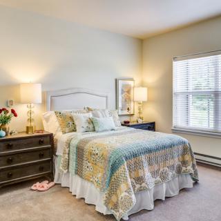 A cozy bedroom with a neatly made bed, featuring a decorative quilt and pillows, flanked by nightstands with lamps. Natural light streams in through a window with blinds.