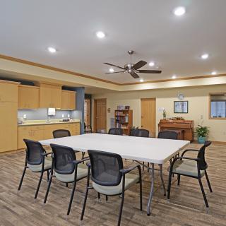 A modern meeting room with a large conference table and chairs, featuring a small kitchen area and elegant wood flooring.