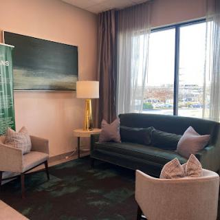 A cozy and modern lounge area featuring a dark green sofa, light-colored armchairs, and large windows that let in natural light.