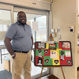 A smiling man stands next to a colorful display board featuring various artwork indoors.