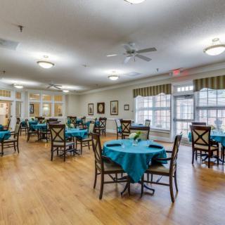 A spacious dining room with multiple tables and chairs, featuring teal tablecloths and large windows allowing natural light.