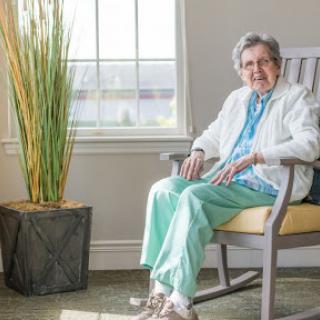 An elderly woman smiles while sitting comfortably in a rocking chair, surrounded by natural light and greenery.