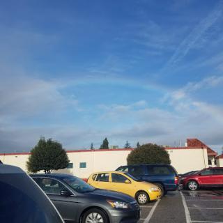 A beautiful rainbow arches across the sky above a parking lot filled with cars.