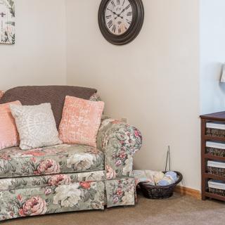 A cozy living room corner featuring a floral sofa with decorative pillows, a wall clock, and a wooden drawer unit.