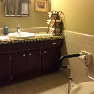 A bathroom featuring a sink and countertop with a decorative towel rack, mirror, and handicap accessible toilet seat.
