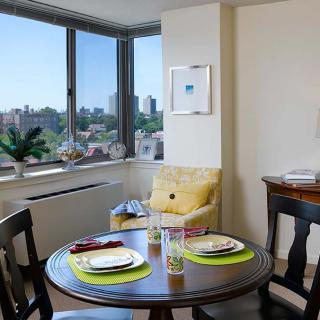 A cozy dining area with a round table set for two, overlooking a cityscape through large windows.