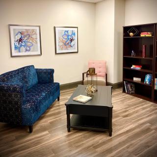 A cozy office with a blue sofa, two framed artworks, and a bookshelf on a wooden floor.
