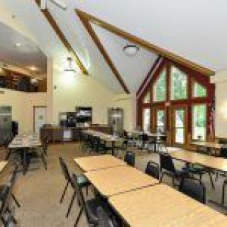 A spacious dining hall with multiple tables and chairs, featuring a high ceiling and large windows.