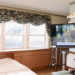 A well-lit hospital room featuring a bed, window with decorative curtains, and a television.