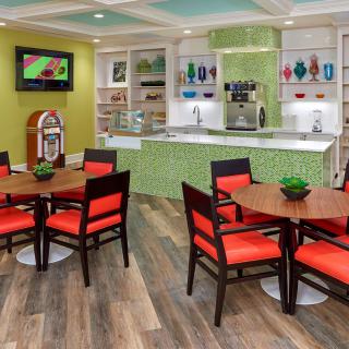 A vibrant cafe interior featuring colorful chairs, round tables, a jukebox, and a counter with various decorations.