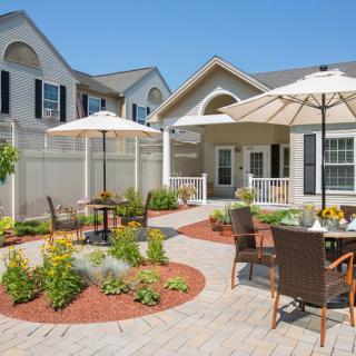 A sunny patio with tables and chairs under umbrellas, surrounded by a beautifully landscaped garden.