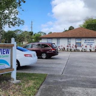 A view of a building with a parking lot, featuring a sign in front and vehicles parked.