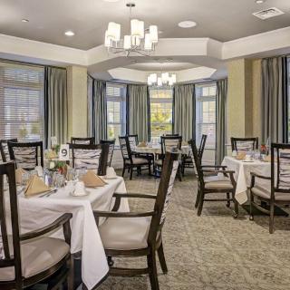 An elegant dining room with neatly set tables and chairs, featuring large windows and soft lighting.