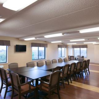 A spacious conference room with a long meeting table surrounded by chairs, featuring large windows and a wall-mounted television.