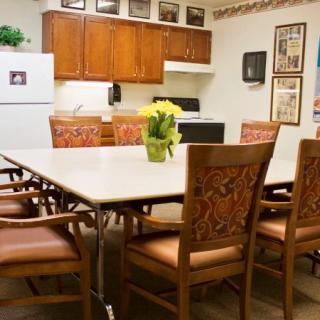 A conference room with a kitchen area featuring a large meeting table surrounded by chairs, decorated with flowers and framed photos on the walls.