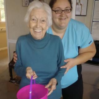 An elderly woman and a younger woman smiling together indoors, holding a pink bowl.