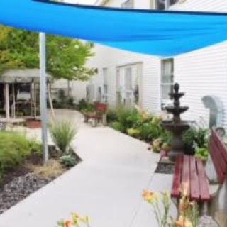 A tranquil outdoor garden area featuring a pathway, benches, a fountain, and a blue shade sail.