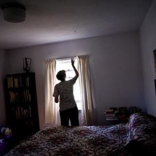 A person standing in a bedroom, drawing back the curtains to let in light.