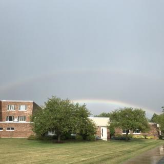 A rainbow arches over a landscape featuring buildings and trees under a cloudy sky.