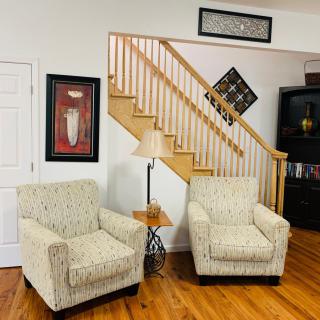 A cozy living room corner with two patterned armchairs, a side table with a lamp, and decorative artwork on the walls near a wooden staircase.