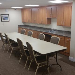 A spacious conference room featuring a long table surrounded by chairs and a kitchenette in the background.