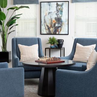 A modern living room with blue chairs centered around a chessboard on a round table, complemented by artwork and an indoor plant.