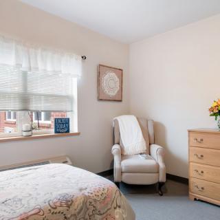 A cozy bedroom with a bed, a comfortable chair, and a dresser next to a window with floral-patterned curtains.