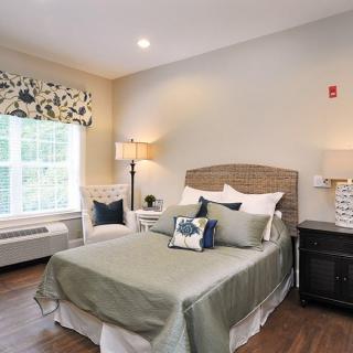 A cozy bedroom interior with a large bed, adorned with pillows and a green throw, flanked by nightstands and lamps. A window with floral curtains allows natural light to brighten the room.