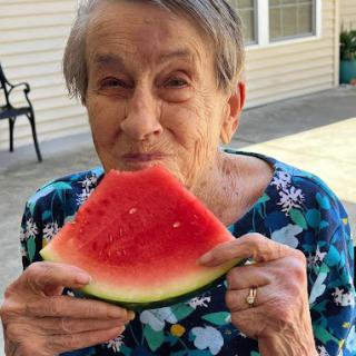 An elderly woman enjoys a slice of watermelon outside on a sunny day.