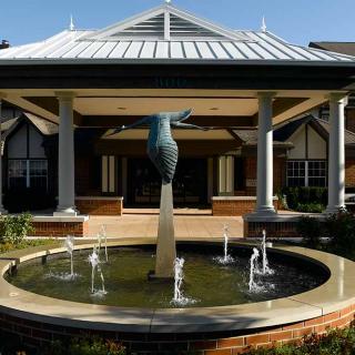 An elegant fountain with a sculpture stands in front of a building with a covered entrance and columns.