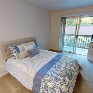 A cozy bedroom featuring a neatly made bed with patterned bedding and pillows, a sliding door leading to a balcony, and soft carpeting.