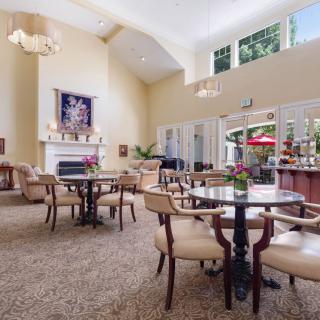 A spacious and elegantly decorated lounge area featuring multiple round tables with chairs, a fireplace, and large windows allowing natural light to flood the room.