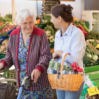 A young woman assists an elderly woman with grocery shopping at a market.