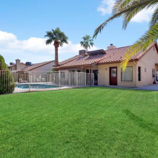 A beautiful house with a spacious backyard featuring a green lawn, swimming pool, and palm trees under a sunny sky.
