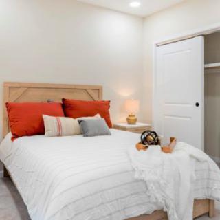 A cozy bedroom with a neatly made bed featuring red and gray pillows, a nightstand with a lamp, and an open closet door.