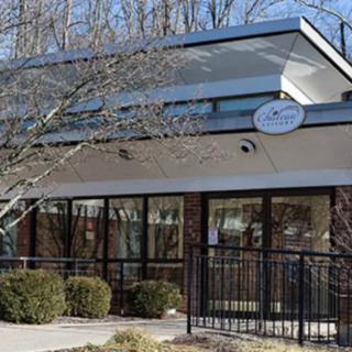 A modern cafe building with large windows and outdoor seating, surrounded by trees.