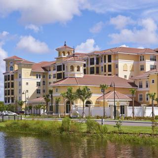 A luxurious residential building with Mediterranean-style architecture, located by a waterfront and surrounded by palm trees.