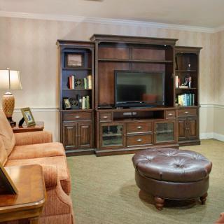A cozy living room featuring an entertainment center with a television, flanked by shelves and cabinets. A comfortable sofa and round ottoman complete the setting.