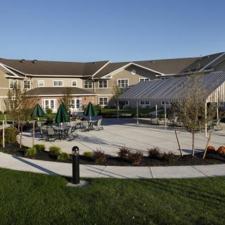 An outdoor area featuring a patio with tables and chairs, surrounded by a landscaped lawn and adjacent to a large building.